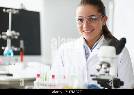 Un employé de laboratoire plutôt gai en blouse médicale examine l'échantillon au microscope Banque D'Images