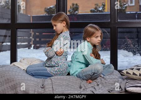 Photo rapprochée de deux petites sœurs mignonnes offensé qui s'assoient sur le dos l'une à l'autre sur le rebord de la fenêtre Banque D'Images