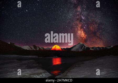 Tente orange lumineuse installée dans un endroit romantique sur la rive d'un lac glaciaire, sur fond de sommets enneigés et d'un sk étoilé Banque D'Images