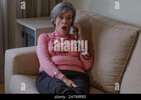 femme mûre allongé sur un canapé faisant un geste surpris tout en ayant une conversation sur son téléphone cellulaire Banque D'Images