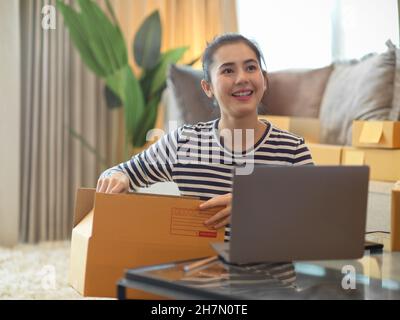 Belle femme entrepreneur préparant des colis et des marchandises d'emballage tout en vérifiant les commandes sur son ordinateur portable. Petite entreprise, e-commerce. Banque D'Images