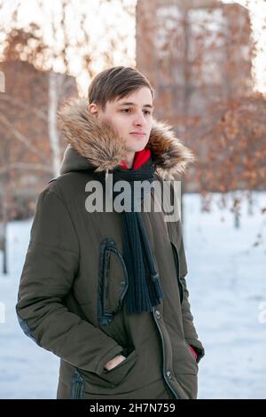 Un jeune homme dans une veste d'hiver à fourrure se dresse dans un parc sur le fond d'une ville enneigée. Banque D'Images