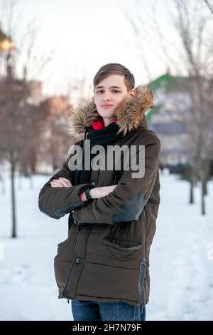 Un jeune homme dans une veste d'hiver à fourrure se tient avec ses bras croisés dans un parc sur le fond d'une ville enneigée. Banque D'Images