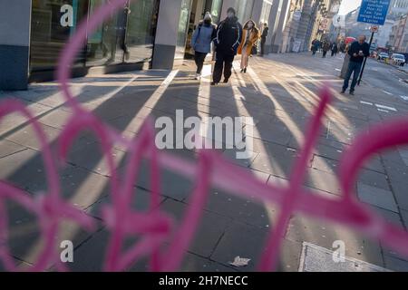 Vu à travers la couleur rose des graffitis sur la fenêtre d'une boîte de téléphone publique, les piétons marchent à travers la lumière solaire d'automne réfléchie sur Moorgate dans la City de Londres, le quartier financier de la capitale, le 23 novembre 2021, à Londres, en Angleterre. Banque D'Images