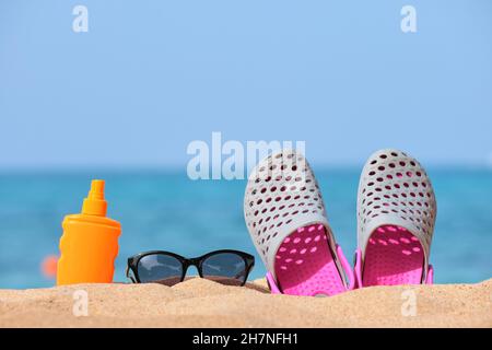 Gros plan sur des chaussures de sabots, de la crème solaire et des lunettes de soleil noires de protection sur une plage de sable au bord de la mer tropicale par temps chaud et ensoleillé. Concept de vacances d'été. Banque D'Images