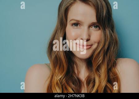 Studio photo de charmante jeune femme timide avec des cheveux bruns ondulés regardant l'appareil photo avec un léger sourire Banque D'Images