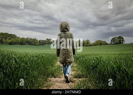 Vue arrière d'une femme portant un parka marchant le long d'une piste dans un champ de blé avec un ciel orageux. Banque D'Images