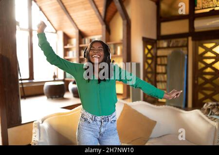 Une jeune femme noire pleine de joie avec un casque sans fil dansant avec les yeux fermés de la musique préférée à la maison Banque D'Images