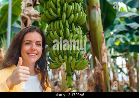 Bonne femme souriante agronome montre approuver le geste de la main positive pouces vers le haut, vérifie le bouquet de bananes fruits de croissance récolte de jeunes palmiers Banque D'Images