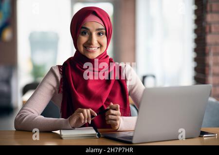 Une femme musulmane joyeuse assise à une table devant un ordinateur portable Banque D'Images