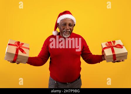 Joyeux homme noir senior en chandail rouge et chapeau de père Noël tenant des boîtes cadeaux de Noël dans les deux mains, offrant de grandes ventes de vacances Banque D'Images