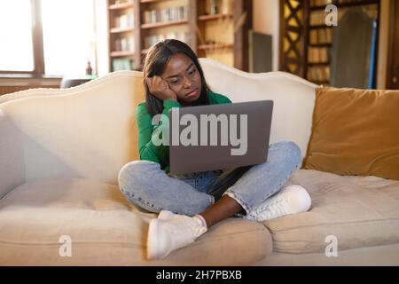 Femme noire fatiguée assise sur un canapé avec un ordinateur portable, regardant à l'écran, se sentant épuisée, ayant un travail en ligne terne à la maison Banque D'Images