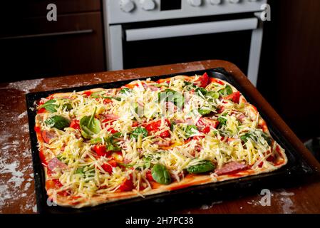 Faire cuire de la pizza avec des saucisses, des tomates, des feuilles vertes en gros plan sur une plaque de cuisson dans la cuisine. Banque D'Images