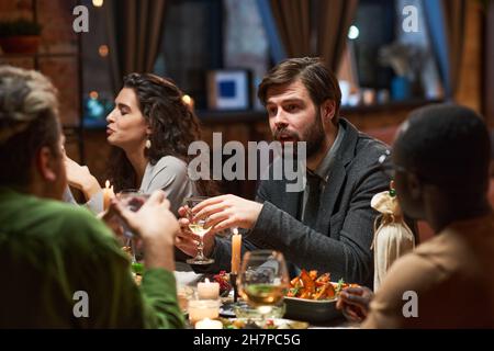 Jeune barbu en costume parlant aux gens tout en étant assis à la table à manger pendant le dîner Banque D'Images