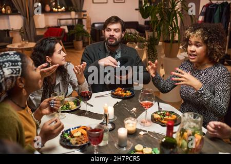 Groupe d'amis assis à la table à manger parlant et jouant à des jeux pendant un dîner Banque D'Images