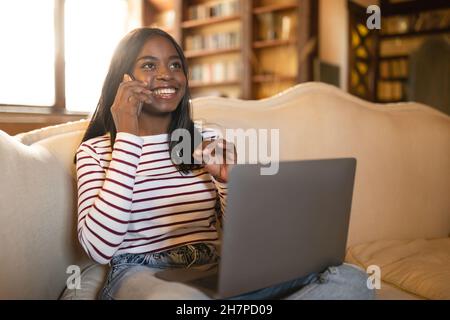 Jeune femme afro-américaine joyeuse ayant une conversation téléphonique, en utilisant un ordinateur portable, assis sur un canapé dans la salle de séjour Banque D'Images