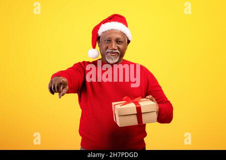 Portrait de l'homme noir senior heureux dans le chapeau du Père Noël tenant un cadeau de Noël, pointant vers vous sur fond orange Banque D'Images