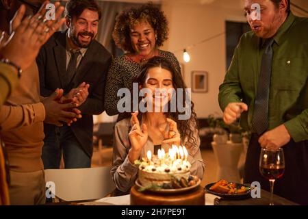 Jeune femme assise à la table avec ses yeux fermés et faisant un souhait avant de souffler les bougies sur le gâteau d'anniversaire Banque D'Images