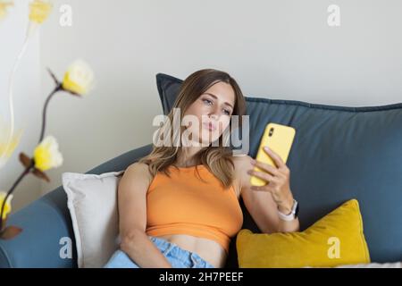 Jeune femme blonde blanche malheureuse se détendant à la maison seule, assis sur un canapé dans une posture confortable, blogueur partageant de bonnes nouvelles sur les médias sociaux via Banque D'Images