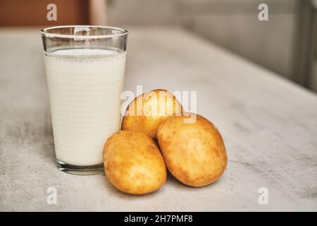 Un verre transparent avec du lait de pomme de terre sur la table à côté des tubercules de pomme de terre. Banque D'Images