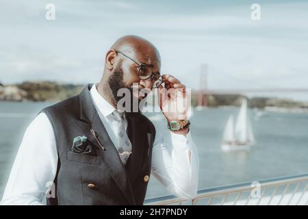Un homme afro-américain de haut niveau avec une barbe noire bien entretenue et dans un costume habillé à la mode avec un gilet ajuste son élégant lunettes de vue par son coup Banque D'Images