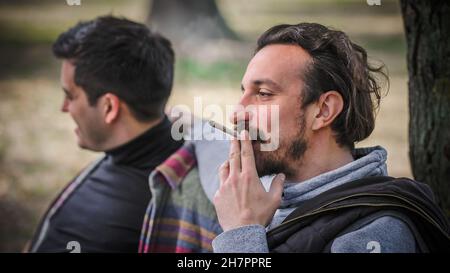 Deux jeunes amis heureux et souriants, qui fument du cannabis, de la ganja ou du hachisch dans un parc public en plein air.Deux hommes fume le thc vert herbe t Banque D'Images