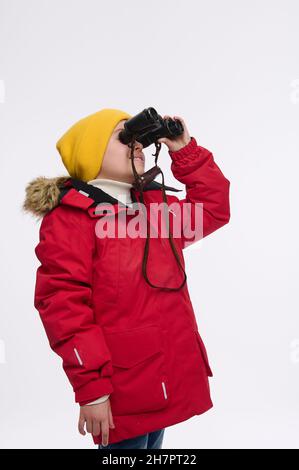 Beau curieux garçon européen d'âge élémentaire dans chapeau jaune et blouson rouge vif, regardant à travers des jumelles, isolé sur fond blanc, copie Banque D'Images