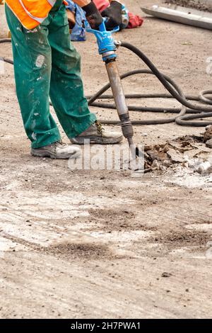 Un ouvrier de la route en combinaison verte éclate de l'asphalte vieux avec un marteau pneumatique sur la route un jour d'été.Image verticale.Copier l'espace. Banque D'Images