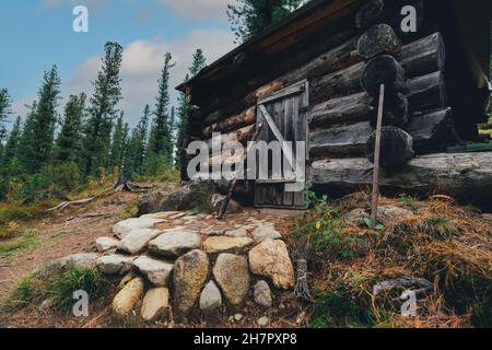 Une vue grand angle d'une cabane de forestier en bois dans une forêt de taïga de conifères profonde sur la glade, construite à partir d'une grande bûche, avec une véranda en pierre sur le terrain cov Banque D'Images