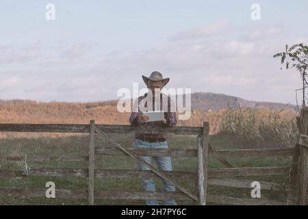 Homme adulte fermier portant chapeau de cow-boy utilisant la tablette pour la communication, la navigation, le shopping en ligne etc.à son champ agricole Banque D'Images