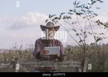 Homme adulte fermier portant chapeau de cow-boy utilisant la tablette pour la communication, la navigation, le shopping en ligne etc.à son champ agricole Banque D'Images