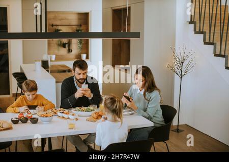 Famille utilisant des téléphones mobiles tout en prenant le petit déjeuner à la table de salle à manger de l'appartement Banque D'Images