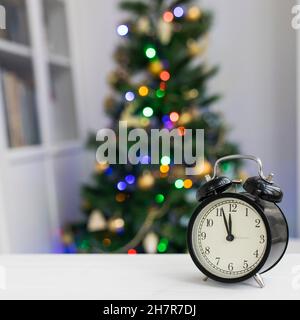 Un réveil noir sur une table blanche indique 12 heures.En arrière-plan, il y a un arbre de Noël hors foyer. Banque D'Images