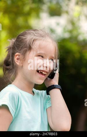 Joyeux enfant d'âge scolaire élémentaire, fille parlant sur son téléphone mobile, tenant un smartphone près de l'oreille portant une montre intelligente, riant.Extérieur Banque D'Images