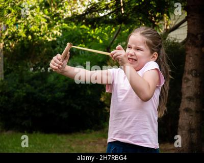 Enfant d'âge scolaire élémentaire, jeune fille tire en arrière un tir de tir de langshot, tirant un rocher faisant un visage drôle, à l'extérieur.Armes jouets, tension et résistance Banque D'Images