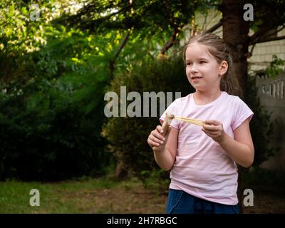 Jeune enfant d'âge scolaire élémentaire, heureuse fille joyeuse tenant un jouet en bois à la langagier dans les mains prêtes à tirer, portrait extérieur, espace de copie.Armes, Banque D'Images