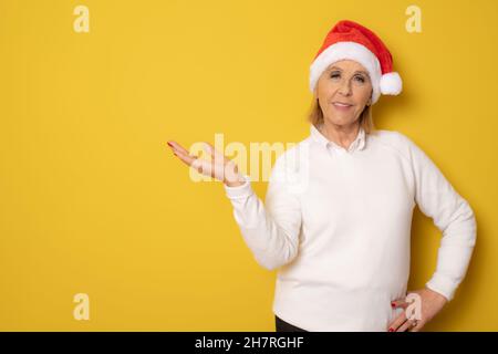 Femme âgée souriante portant un chapeau de père noël montrant le produit avec la main isolée sur fond jaune. Banque D'Images