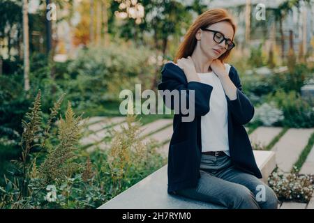 La prise de vue extérieure d'une femme à tête rouge pose des douleurs aux épaules à l'extérieur dans le parc pendant la journée Banque D'Images