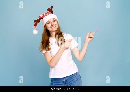 Gros plan portrait, elle est belle, charmante petite fille gaie avec des cornes de cerf en t-shirt blanc, sauvage look festif, montre un signe avec ses doigts sur le côté sur un fond bleu. Banque D'Images