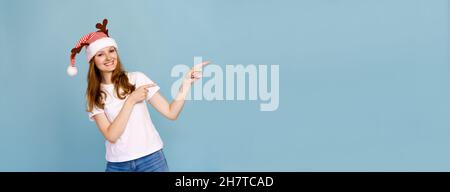 Gros plan portrait, elle est belle, charmante petite fille gaie avec des cornes de cerf en t-shirt blanc, sauvage look festif, montre un signe avec ses doigts sur le côté sur un fond bleu. Banque D'Images