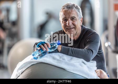Un homme âgé qui se repose sur une balle d'exercice dans la salle de gym tient une bouteille d'eau en regardant l'appareil photo. Banque D'Images