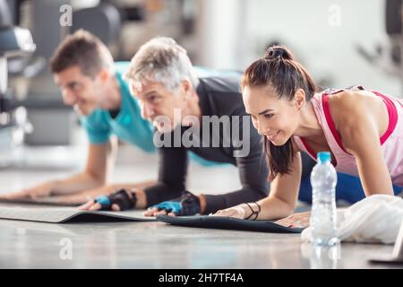 Groupe d'âge divers faisant des planches de coude sur les tapis à l'intérieur de la salle de gym avec une bouteille d'eau en plastique à l'avant. Banque D'Images