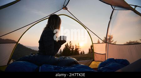 Matin dans le camp de tente.Silhouette de voyageur solitaire, assis dans une tente, tenant une tasse chaude dans ses mains, et regardant le lever du soleil sur les collines de montagne. Banque D'Images