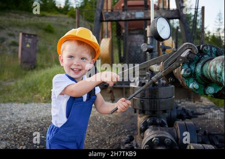 Adorable petit garçon dans le casque de construction à l'aide d'une clé industrielle lors de la réparation du tuyau de cric de pompe.Adorable enfant dans une combinaison de travail regardant de côté et souriant tout en serrant le boulon sur le tuyau. Banque D'Images