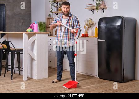 Jeune homme beau barbu dans la cuisine, portant une chemise à carreaux, essaie de faire face à une pelle et un balai. Banque D'Images