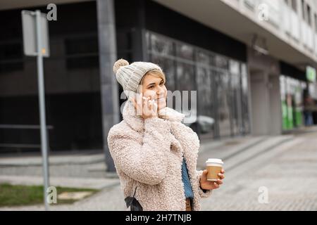 Portrait de femme mignonne en bonnet tricoté et manteau de fourrure en fausse fourrure avec une tasse de café chaud dans sa main parlant par téléphone mobile, style de rue, moderne Banque D'Images