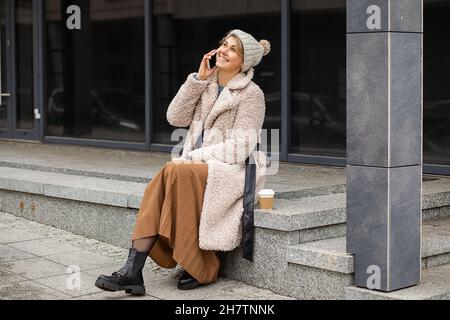 Jeune femme mignonne avec un café dans une tasse jetable, utilisant un téléphone portable, assis souriant sur l'escalier de la rue européenne, portant un faux manteau de fourrure et un chapeau, St Banque D'Images