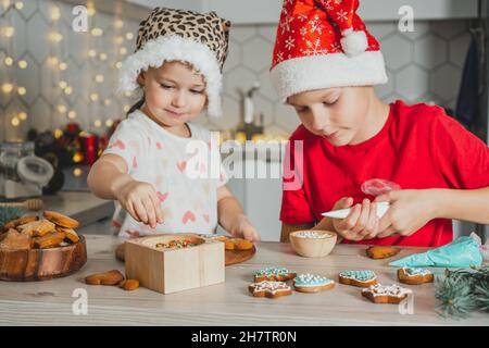 Petite fille à poil foncé de 3 ans et garçon de 8 ans en chapeau de père Noël rouge décorent les biscuits au pain d'épice avec du glaçage.Les frères et sœurs décorent Noël blanc Banque D'Images