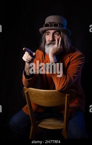 Un intellectuel barbu à cheveux gris d'âge mûr dans un chapeau de steampunk et une veste orange gestuelle soigneusement, pointant vers un interlocuteur invisible Banque D'Images