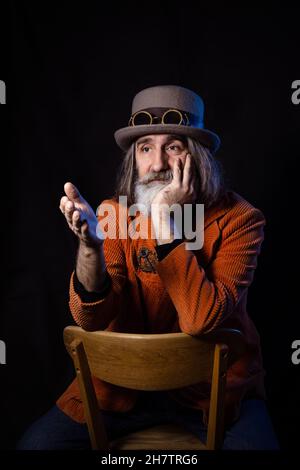 Un intellectuel barbu à cheveux gris d'âge mûr dans un chapeau de steampunk et une veste orange gestuelle soigneusement, pointant vers un interlocuteur invisible Banque D'Images